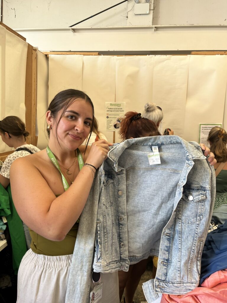 photo of inside a warehouse for our Kilo Sale and a lady wearing a green vest is holding up a light blue denim jacket to show it off with a pile of clothes behind her