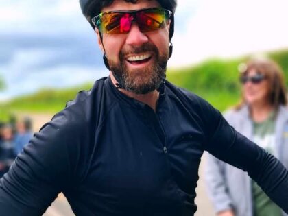 a photo of a man in a black cycling outfit with a helmtet and glasses and greenery behind him