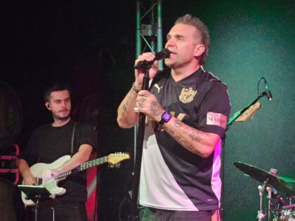 a dimly lit stage with a robbie williams impersonator singing and a man with a guitar and a drum kit behind him