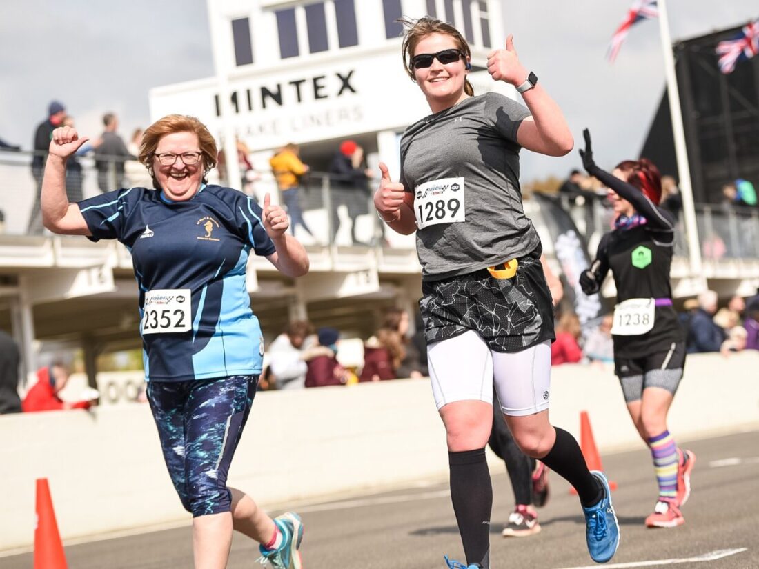 Two women running and smiling to the camera as they run past. To their left are barriers and people cheering. The woman on the left is shorter and wears a blue t-shirts and blue 3/4 length leggings, the woman on the right wears a grey t-shirt and double layered grey and white shorts.