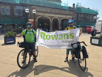 a photo of two men wearing green rowans hospice t shirts on their bikes, holding a white rowans hospice banner between them infront of a building in the sunshine