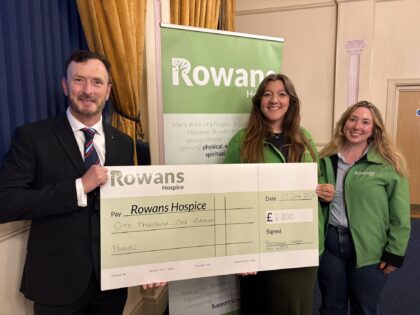 photo of two girls in green rowans jackets holding a cheque with a man in a suit in a room with rowans hospice banners behind them