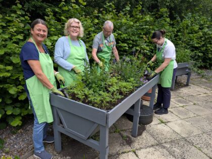 a photo of 4 people around a garden display which they are putting plants into with gloves and a green rowans hospice apron