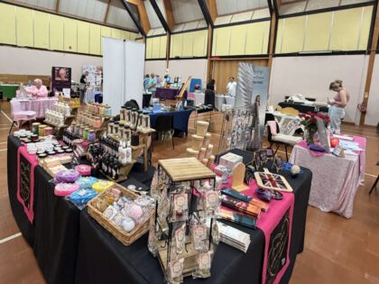 a sportshall room with multiple stalls full of colorful jewellery, offering bath soaps and reiki with people browsing around