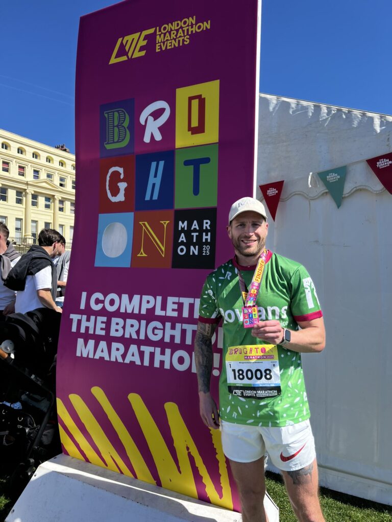Man smiling and holing a brighton marathon medal, next to a banner saying "I completed the Brighton Marathon"