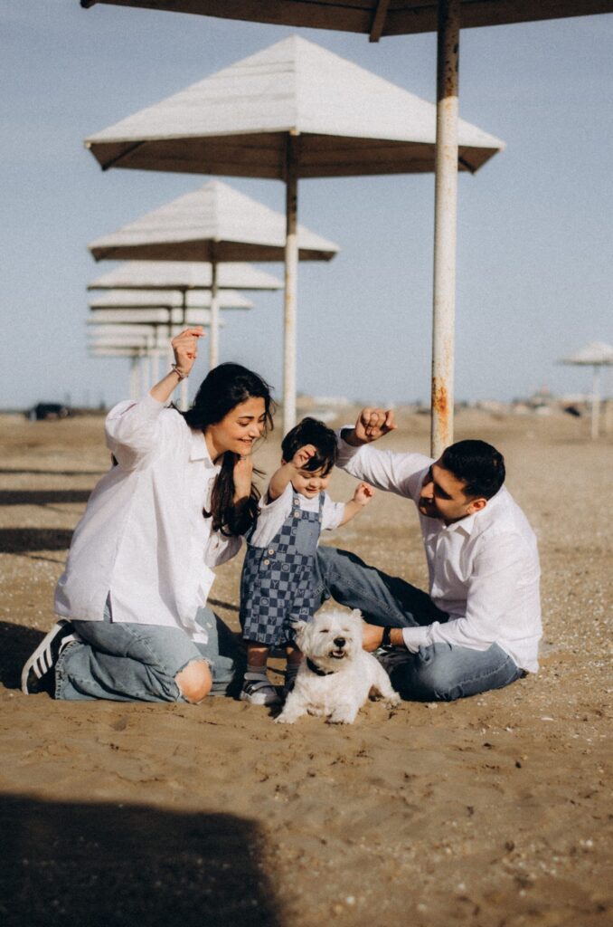 picture of family at the beach having fun. the mum is to the left, child in the middle and dad to the right, in front of the child is a small white dog and they all look at him