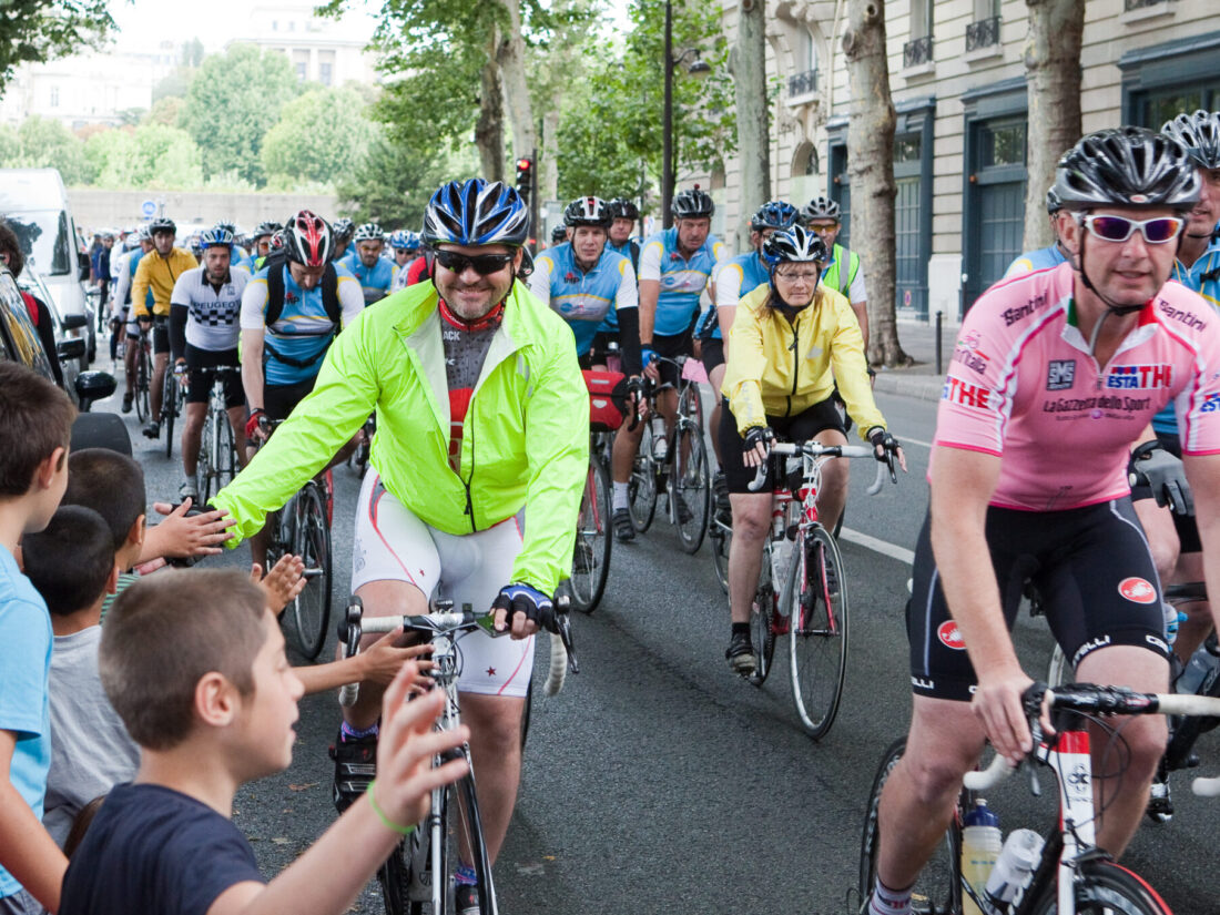 Man on a bicycle, wearing a helmet and a bright green jacket, his hand is out as he high-fives people watching. Around him are other cyclists