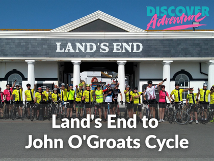 Group of cyclists in gear, holding their bikes to the side of them, in bront of building with "Lands End" acorss. There is text over the image "Land's End to John O'groats Cycle", you can also see a pink and blue logo on the top right corener of the pictures that says "Discover Adventure"