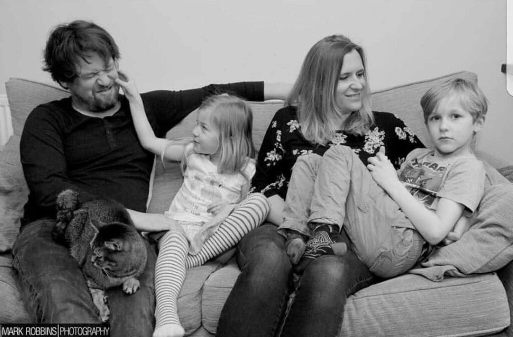 Black-and-white family photograph showing two adults and two children sitting together on a sofa with a cat. The young girl reaches up to touch the man’s face, while the boy sits beside the woman with his arms folded.