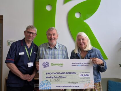photo of two people and a male nurse holding a cheque saying prize winner, stood in a room infront of a large green R from the rowans hospice logo