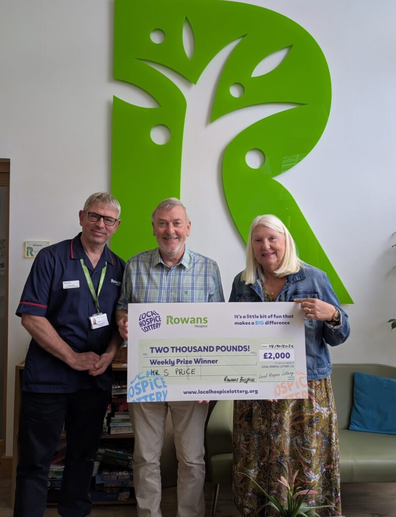 photo of two people and a male nurse holding a cheque saying prize winner, stood in a room infront of a large green R from the rowans hospice logo